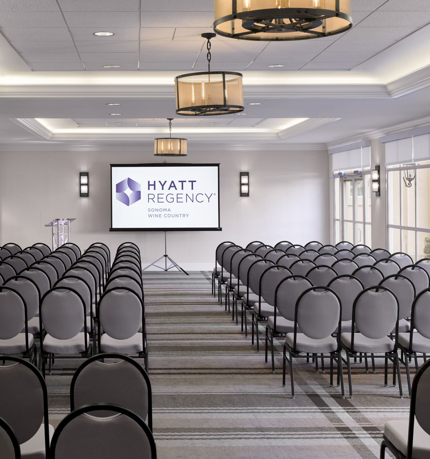 A conference room with rows of chairs facing a screen displaying the Hyatt Regency logo, featuring large windows, lighting fixtures, and a podium.