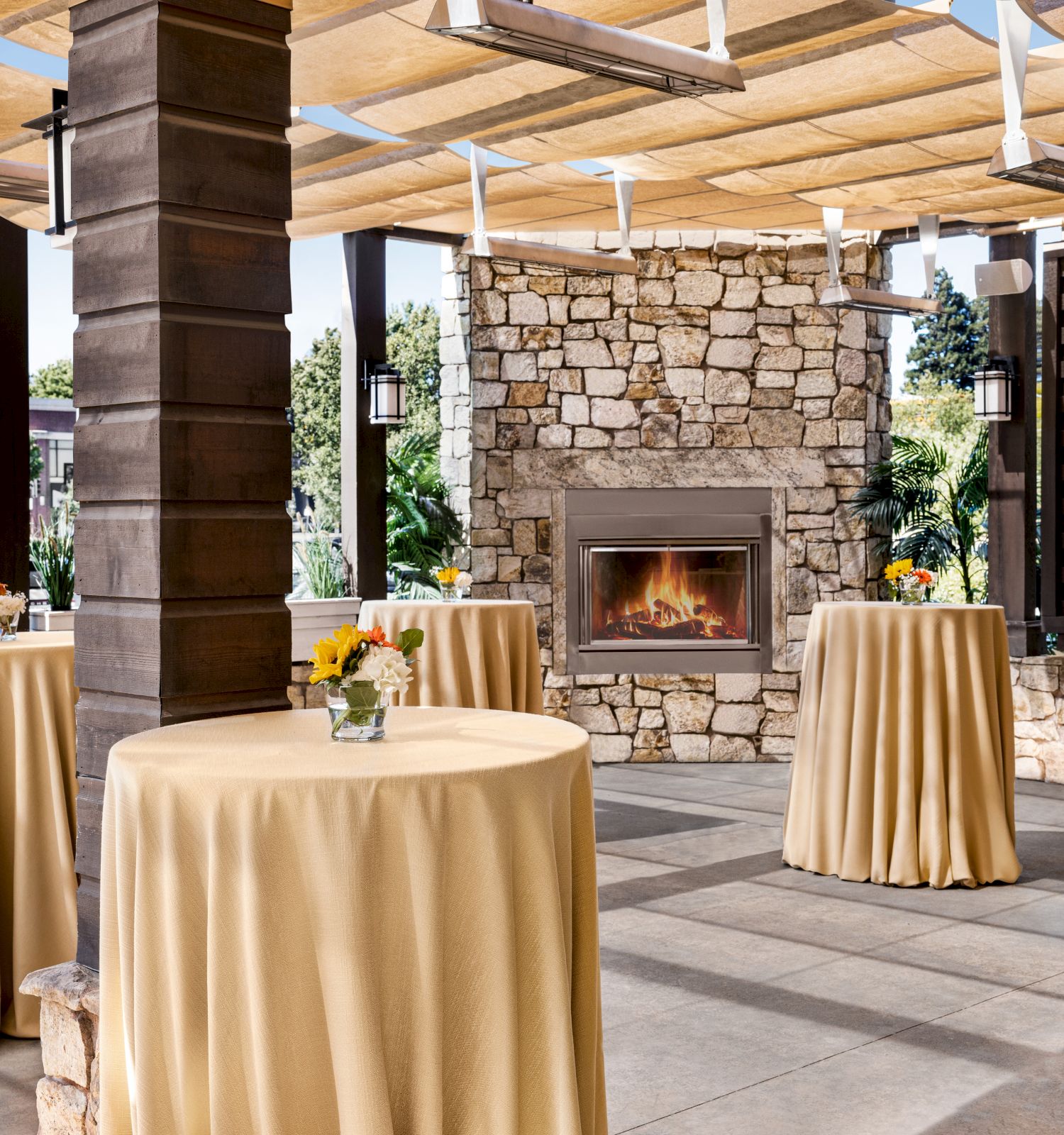 An outdoor event space with round tables draped in beige cloths and topped with small floral centerpieces, featuring a stone fireplace.