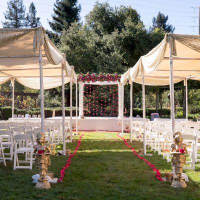 An outdoor wedding setup with white chairs, ceremonial statues, flower arrangements, and a decorated stage under a canopy on a grassy area.