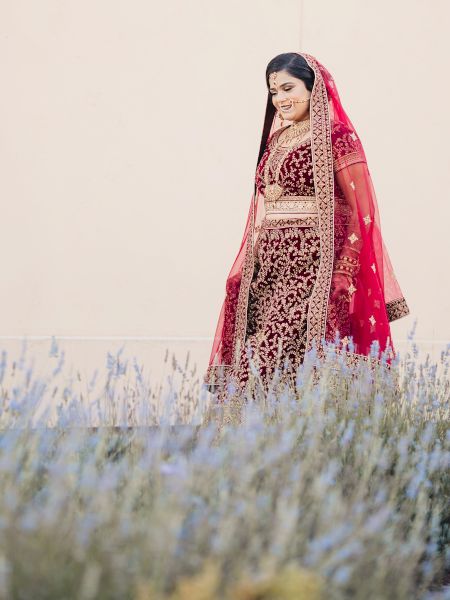 A person wearing a traditional red and gold outfit, standing in front of a beige wall with lavender bushes in the foreground.