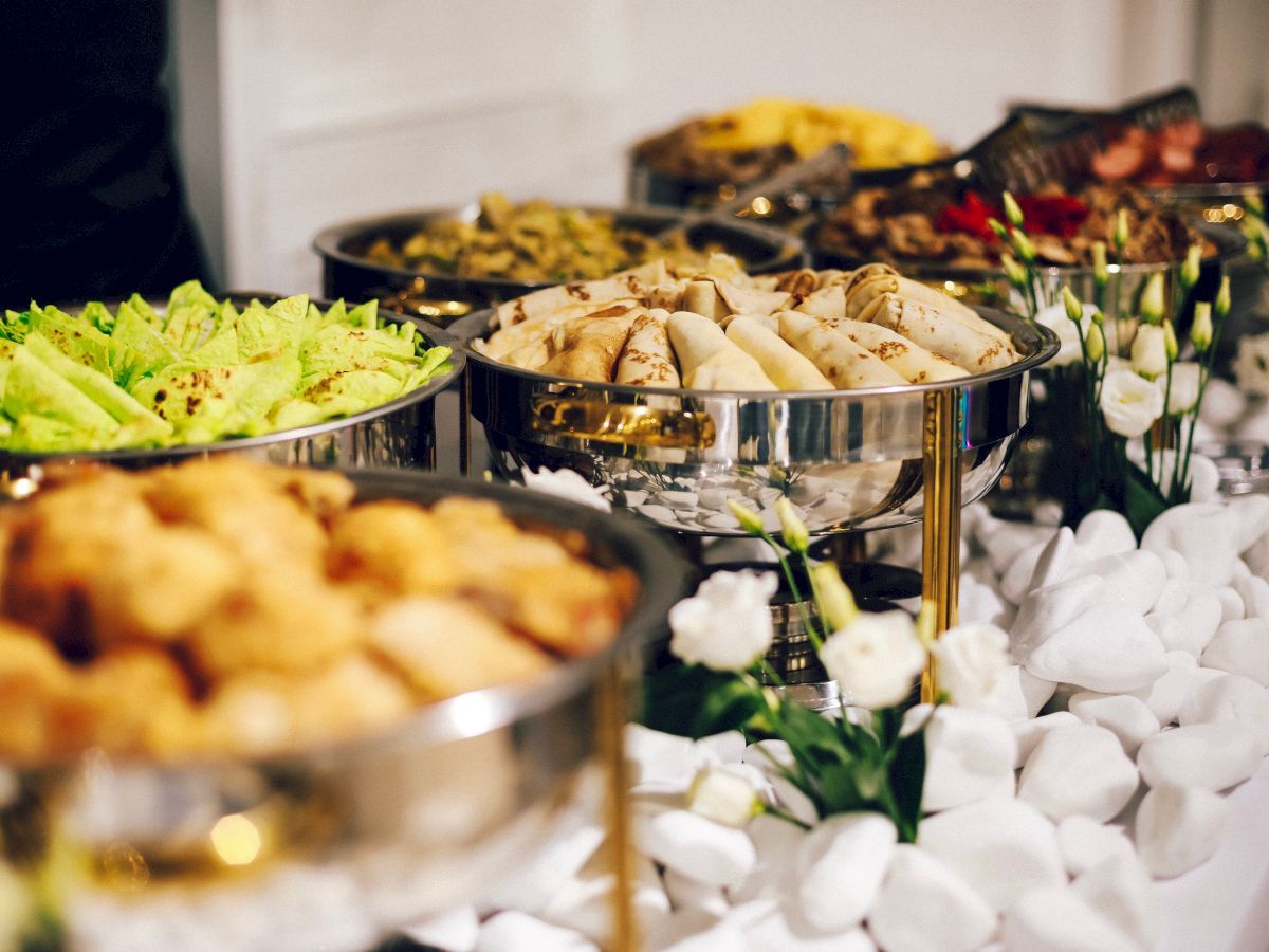 A buffet setup with a variety of dishes in stainless steel chafing dishes, surrounded by decorative white pebbles and flowers, is shown.