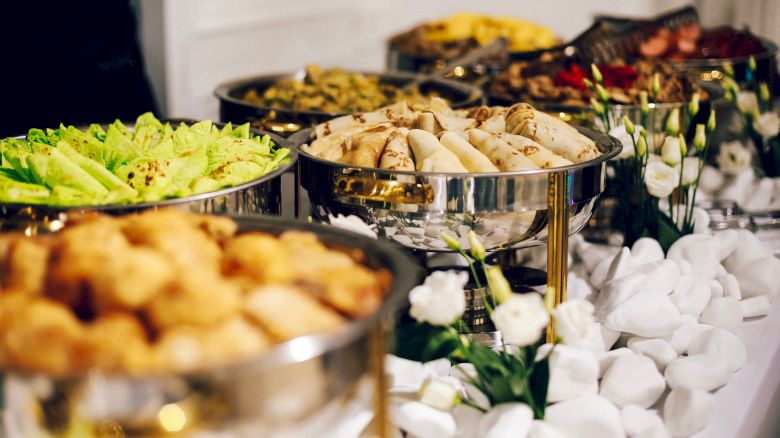 A buffet setup with a variety of dishes in stainless steel chafing dishes, surrounded by decorative white pebbles and flowers, is shown.