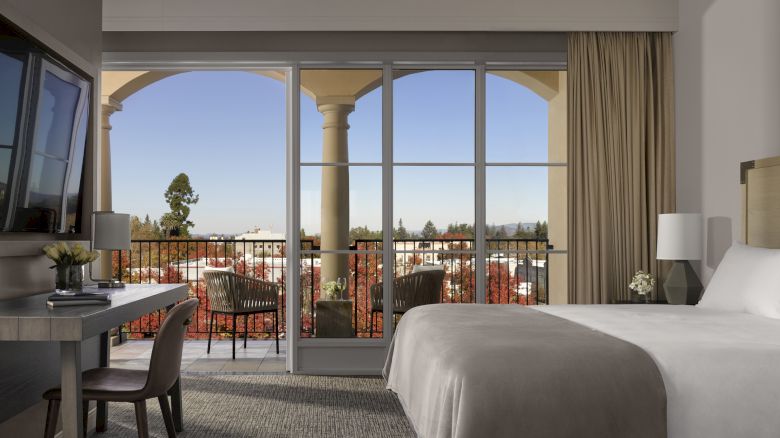 A cozy bedroom featuring a large bed, desk with TV, and a balcony with chairs overlooking a scenic outdoor view, framed by arching columns.