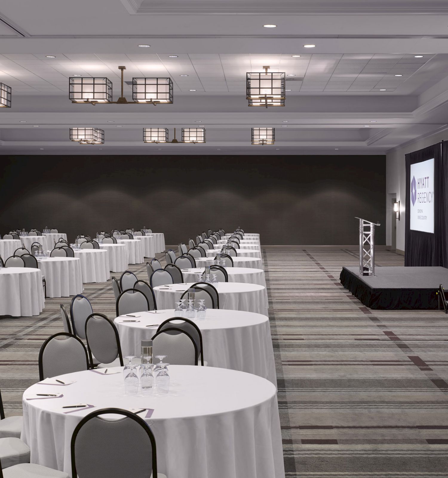 A conference room setup with round tables and chairs, a podium on a stage, and large screens displaying the 