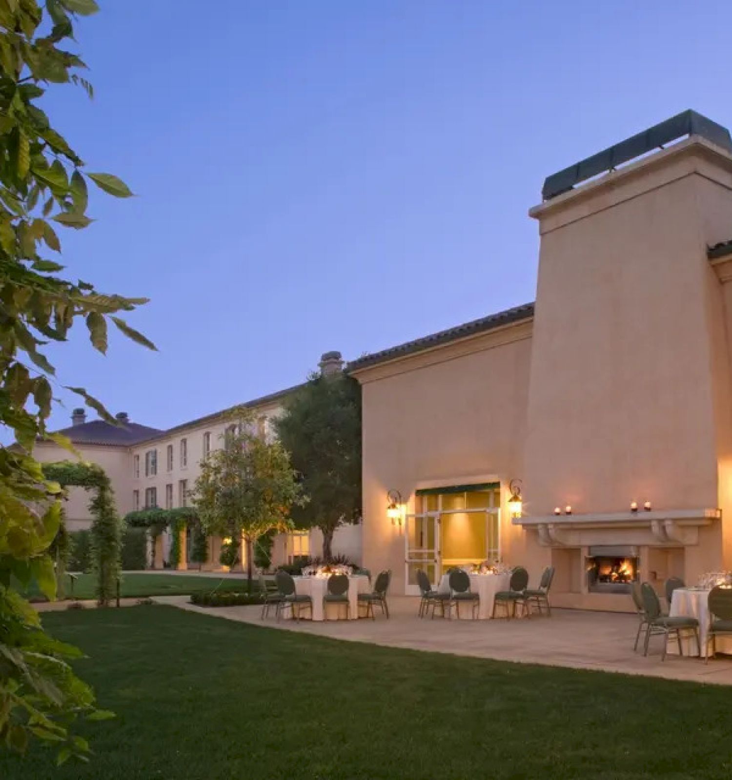 The image shows an outdoor patio with tables, chairs, and a fireplace surrounded by greenery. Buildings and trees are visible in the background.