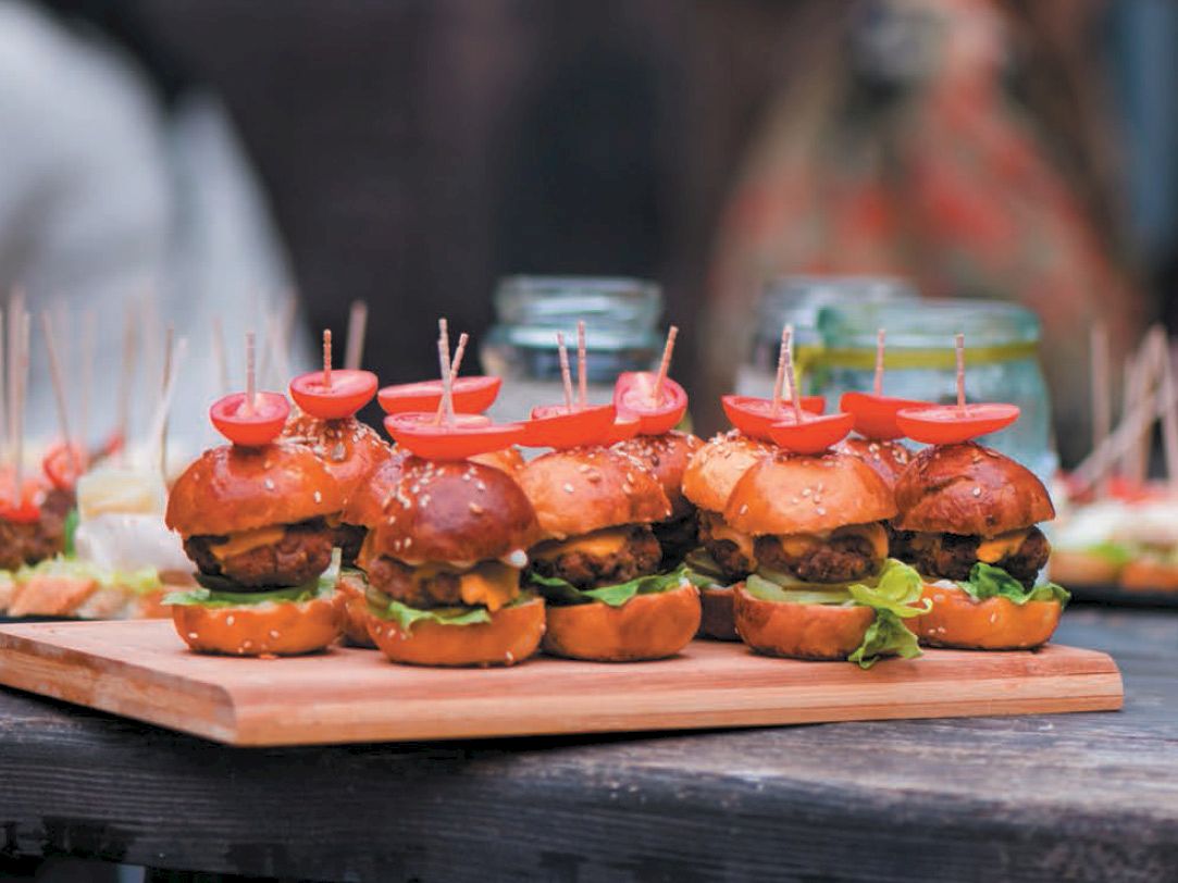 Sliders on a wooden board, topped with cocktail sticks and cherry tomatoes, displayed on a wooden table with blurred food in the background.