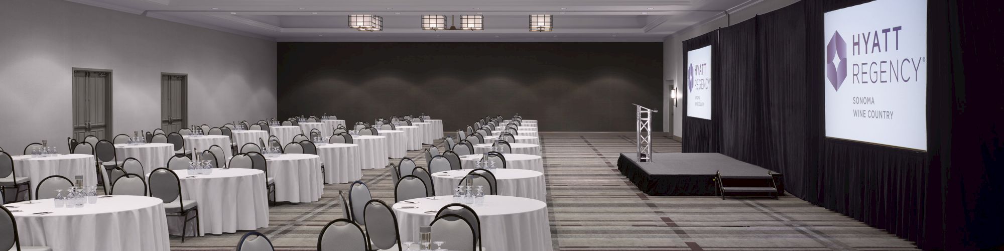 A large conference room set up with round tables, chairs, and a stage with presentation screens. The screens display 