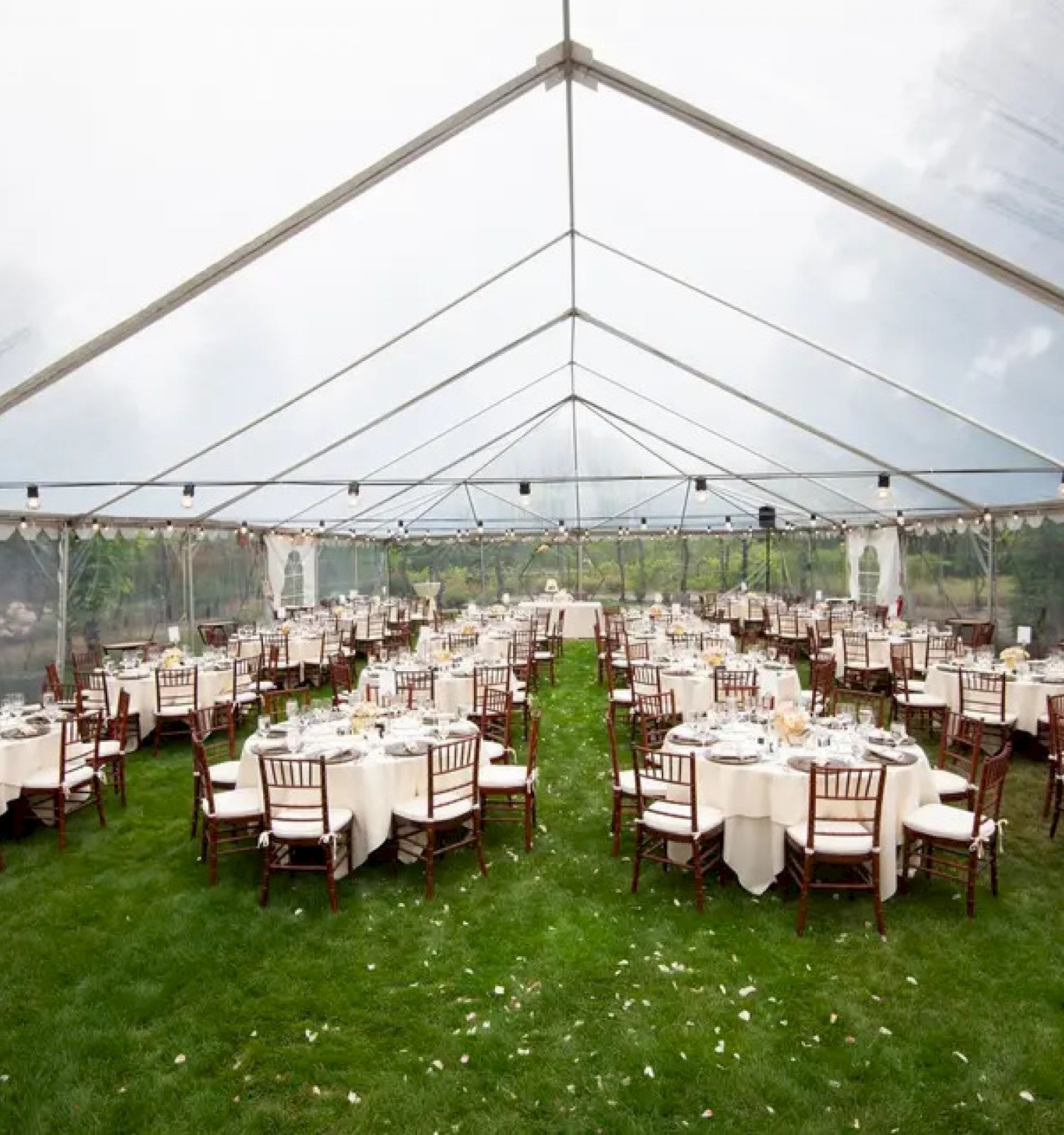 The image shows an outdoor event setup under a large transparent tent with round tables and chairs arranged on a grass lawn, prepared for a gathering.