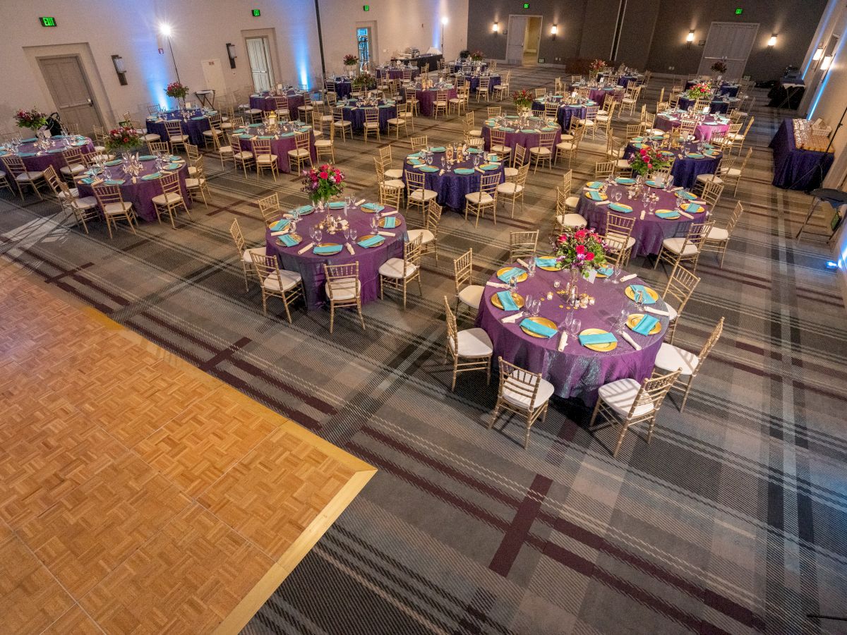 A banquet hall with neatly arranged tables, purple tablecloths, and elegant place settings, set for an event with a dance floor on the side.