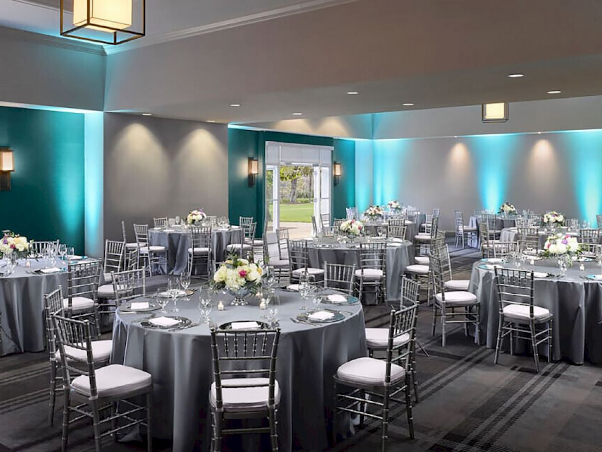 The image shows an elegant banquet hall with round tables set for an event. The tables have gray tablecloths and centerpieces with flowers.