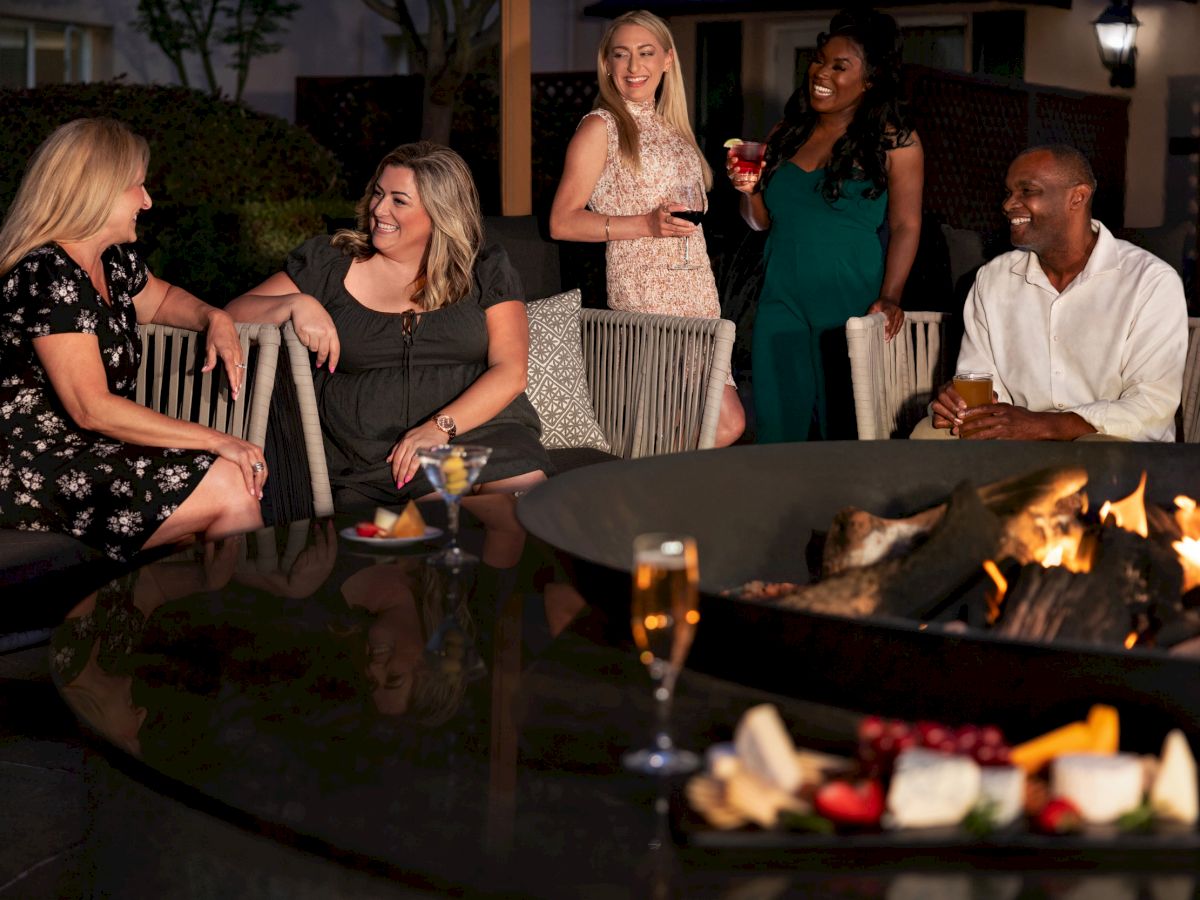 A group of people are sitting and standing around an outdoor fire pit, enjoying drinks and snacks in a relaxed, social setting.