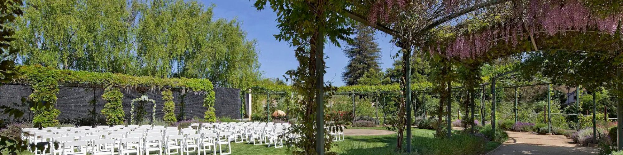 An outdoor wedding setup with rows of white chairs facing an altar, surrounded by greenery and a pathway covered with flowering vines.