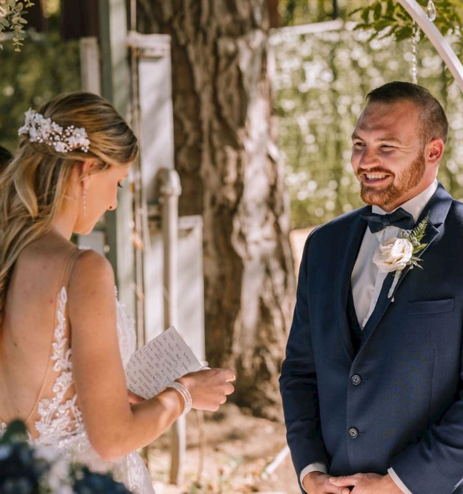 A couple is getting married outdoors; the bride is reading vows while the groom smiles. There are also two people in the background.