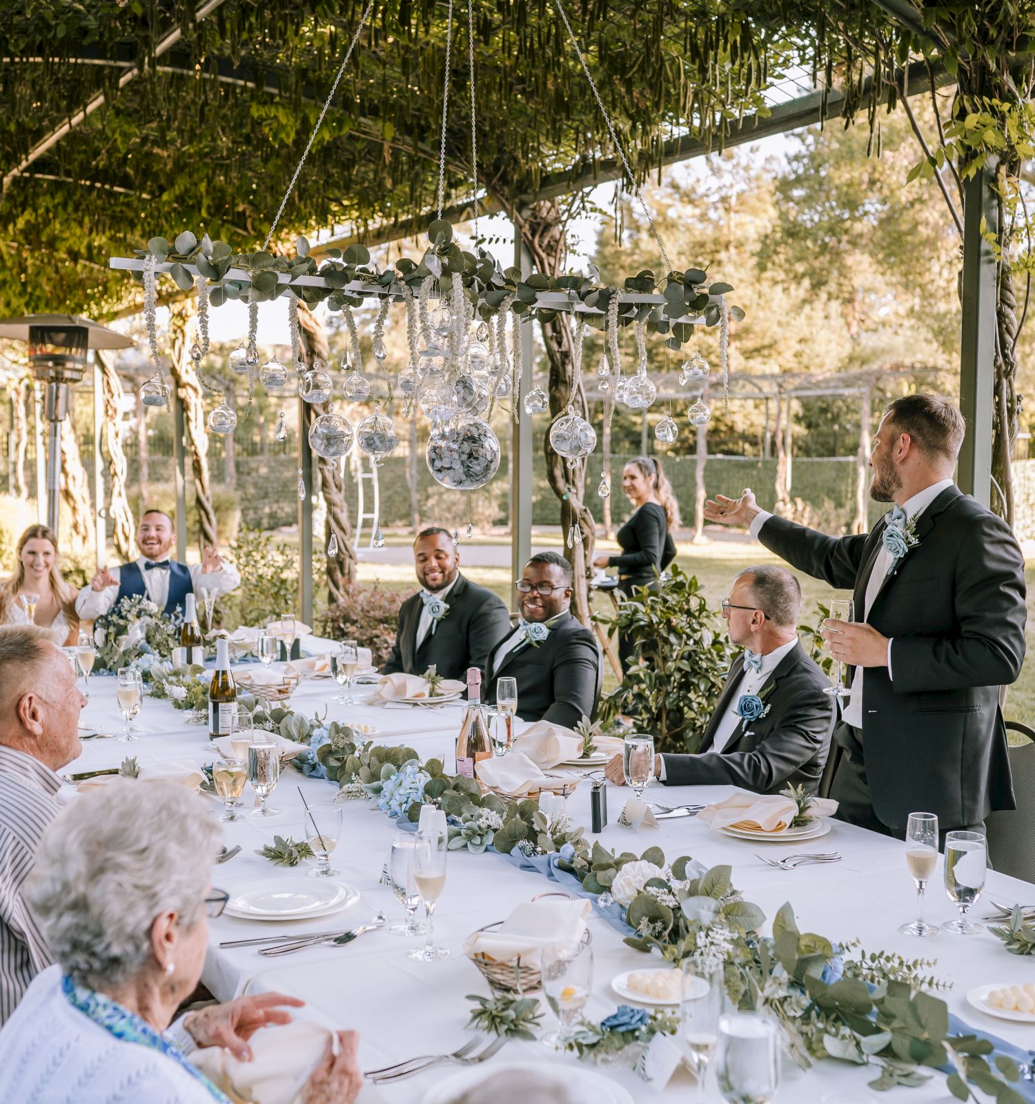 People are gathered around a long outdoor dining table, with one man giving a toast. The setting is decorated with greenery and hanging lights.