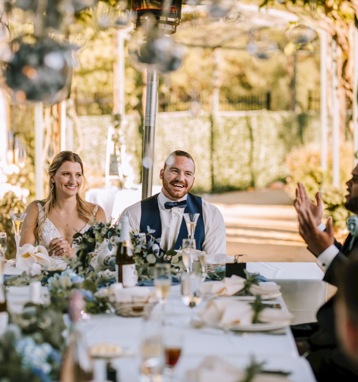 A bride and groom sit at a decorated table during an outdoor wedding reception, smiling and conversing with guests.