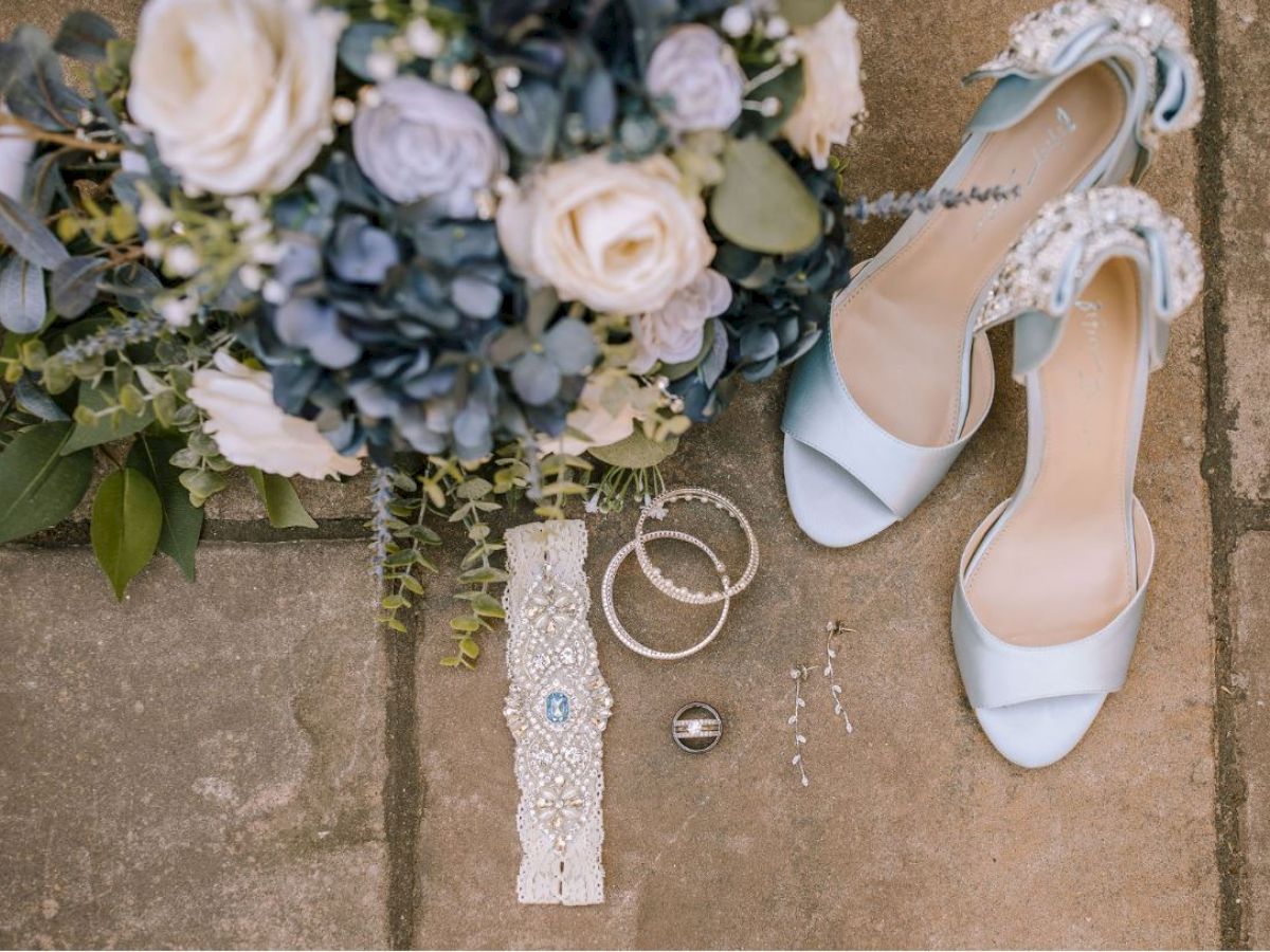 A bouquet, blue high heels, jewelry, a garter, and a wedding ring are arranged on a stone floor, possibly for a wedding.