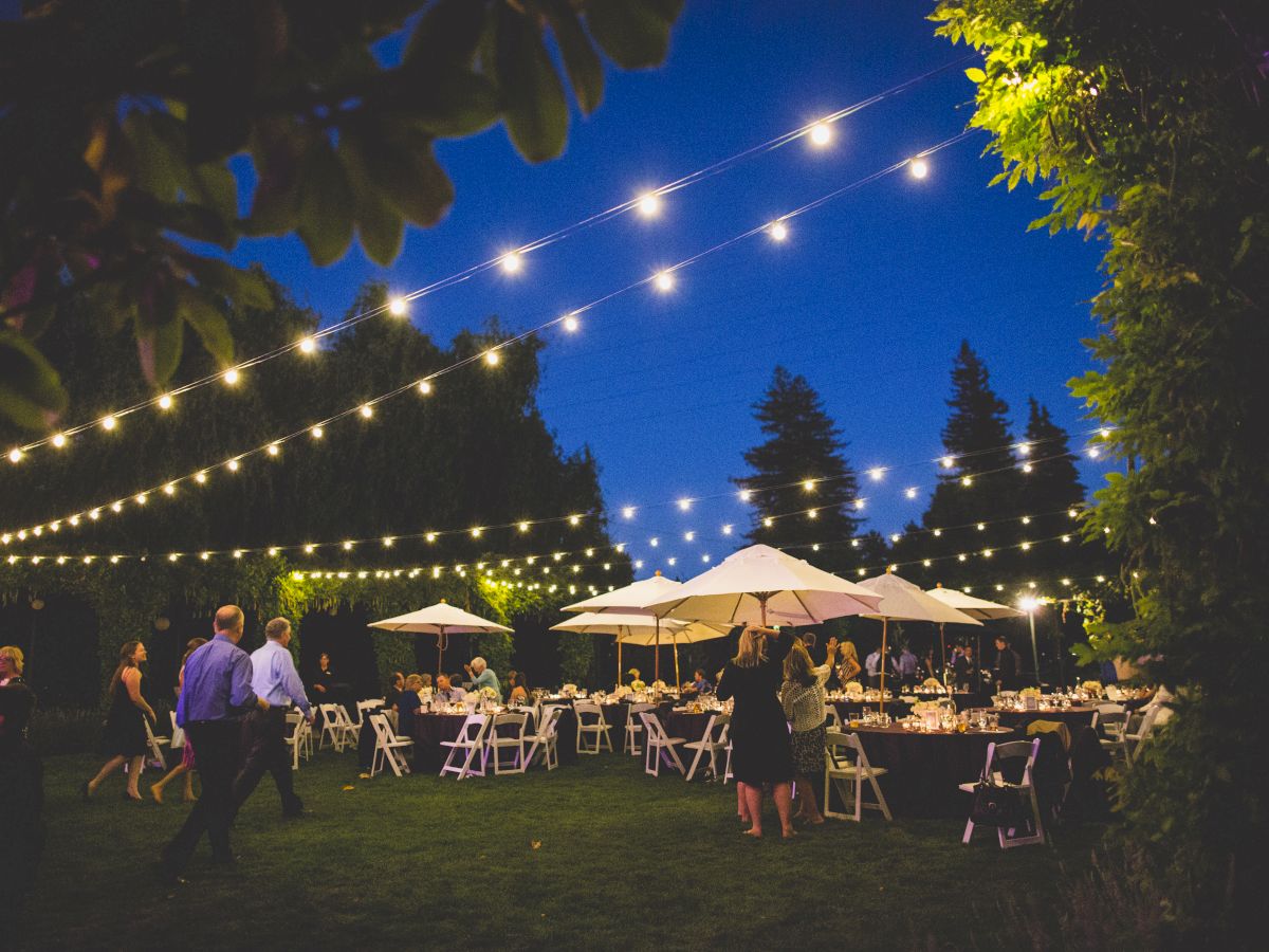 An outdoor evening event with string lights, tables, chairs, and umbrellas, attended by people socializing under a clear night sky, is depicted.