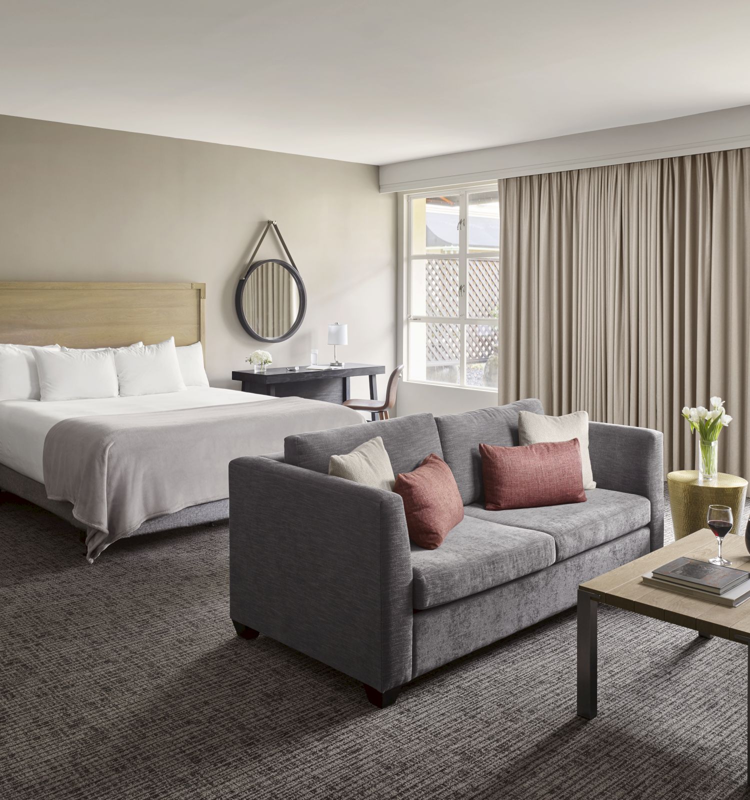 This image shows a well-furnished hotel room with a bed, seating area, and natural light from a window with curtains.