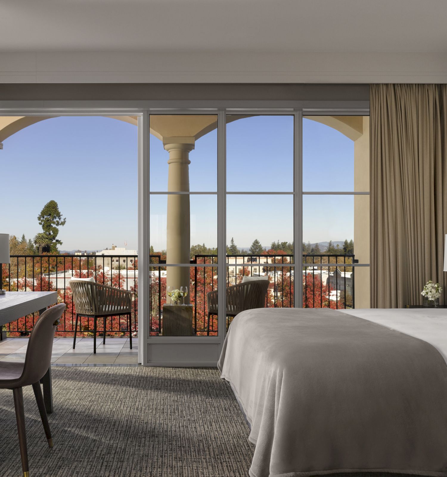 A modern hotel room with a bed, desk, TV, and a balcony with a view of a cityscape, featuring arches and trees in the background.