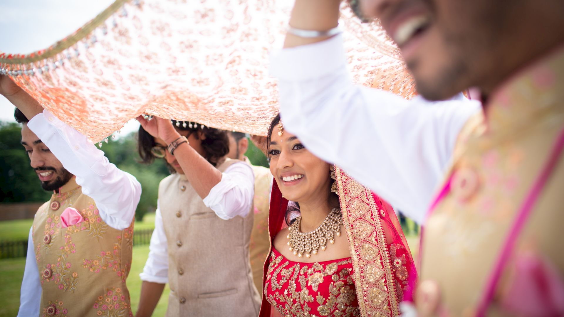 A bride is walking under a decorative cloth held by four people, likely during a wedding ceremony, and everyone appears happy and celebratory.