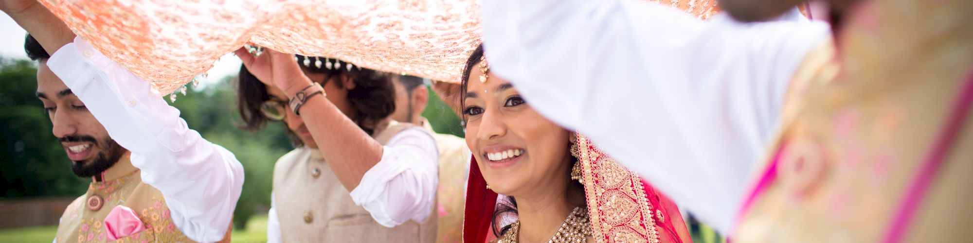 A bride is walking under a decorative cloth held by four people, likely during a wedding ceremony, and everyone appears happy and celebratory.