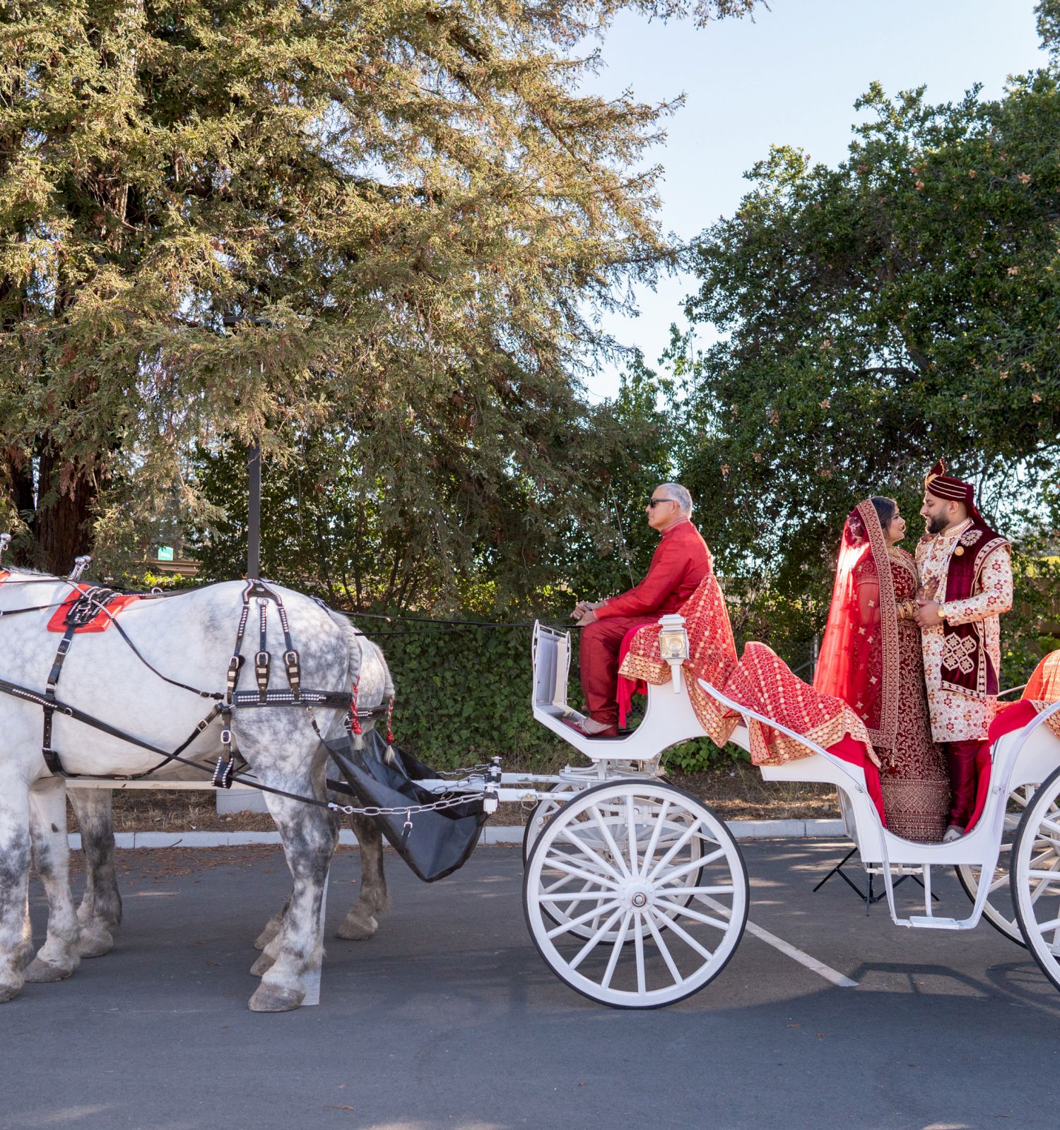 A decorated white horse-drawn carriage with two people in traditional attire is parked in an outdoor setting with trees in the background.