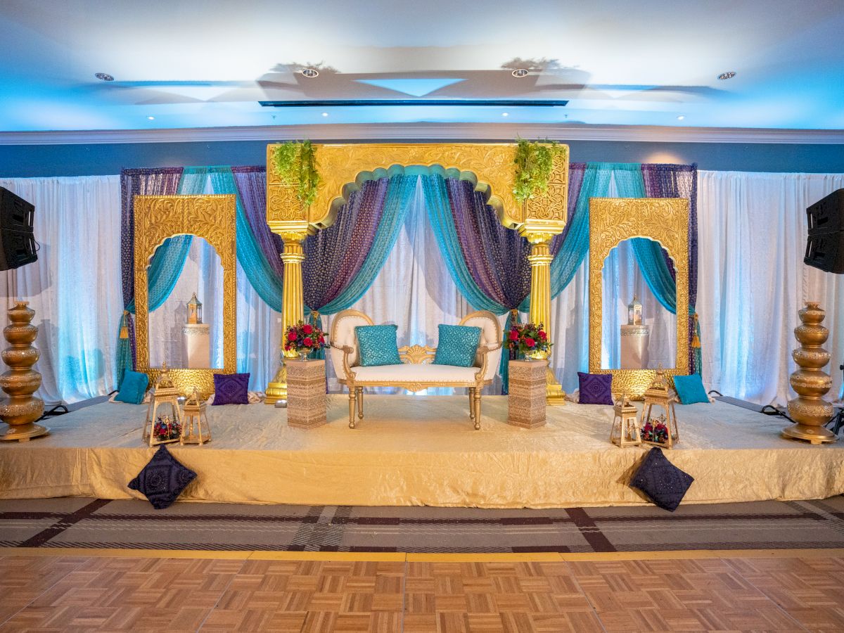 A beautifully decorated stage with luxurious drapes, a white and gold seating area, blue and gold pillows, flanked by ornate columns and floral arrangements.