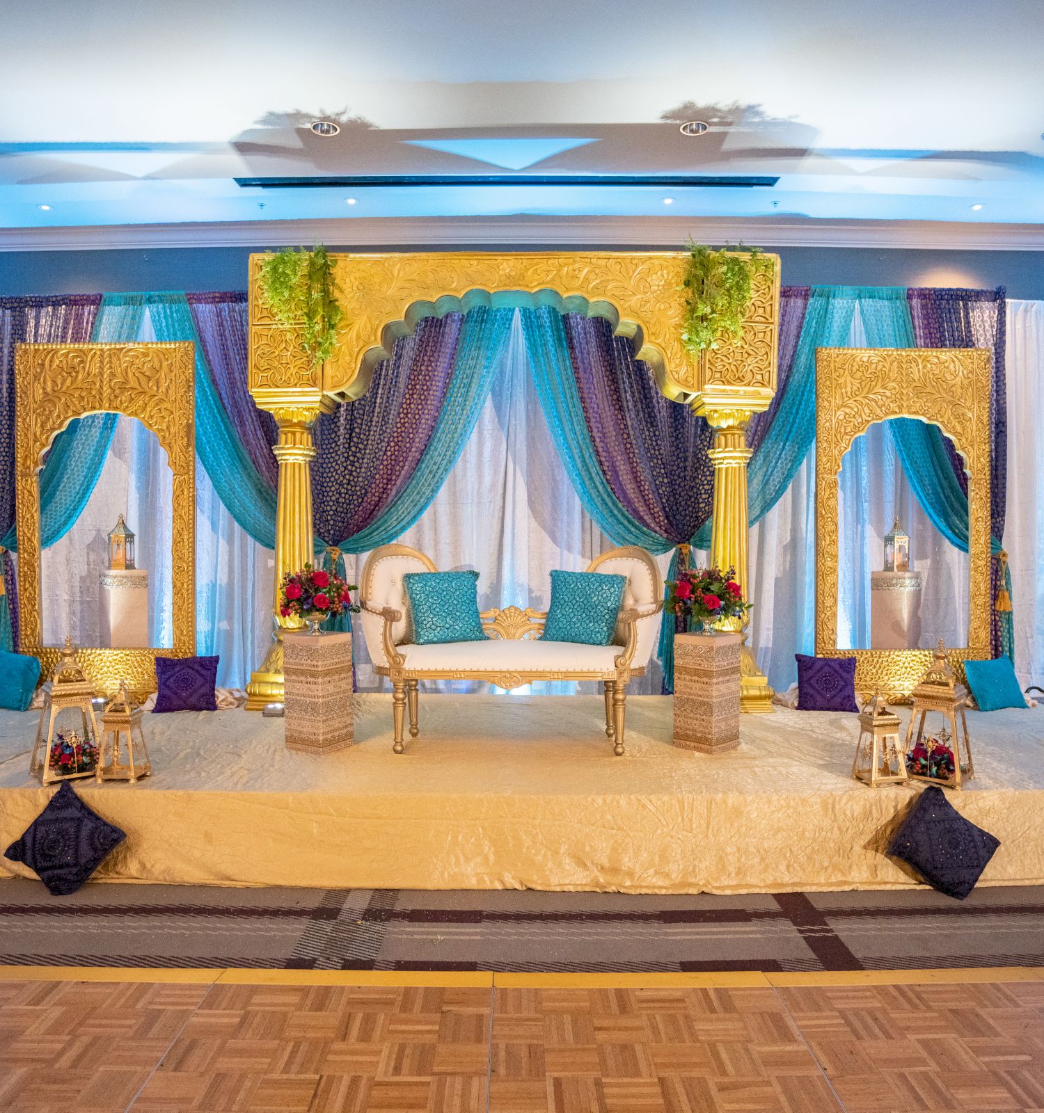 An elegant stage setup with a gold and teal theme, featuring a central white couch, flanked by decorative pillars and draped with fabric.