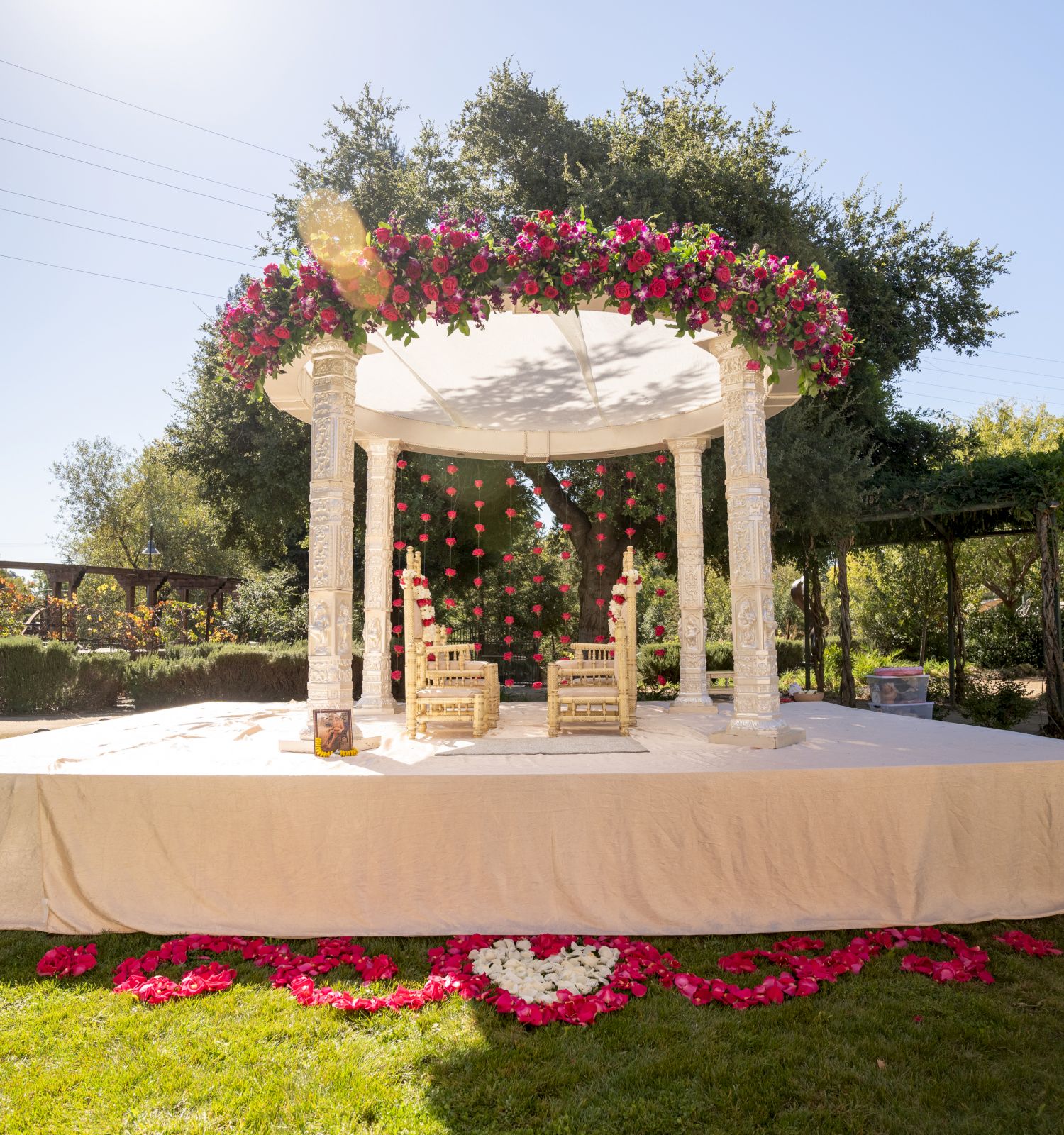 A beautifully decorated outdoor wedding mandap with two ornate chairs under a floral canopy, set in a lush garden.