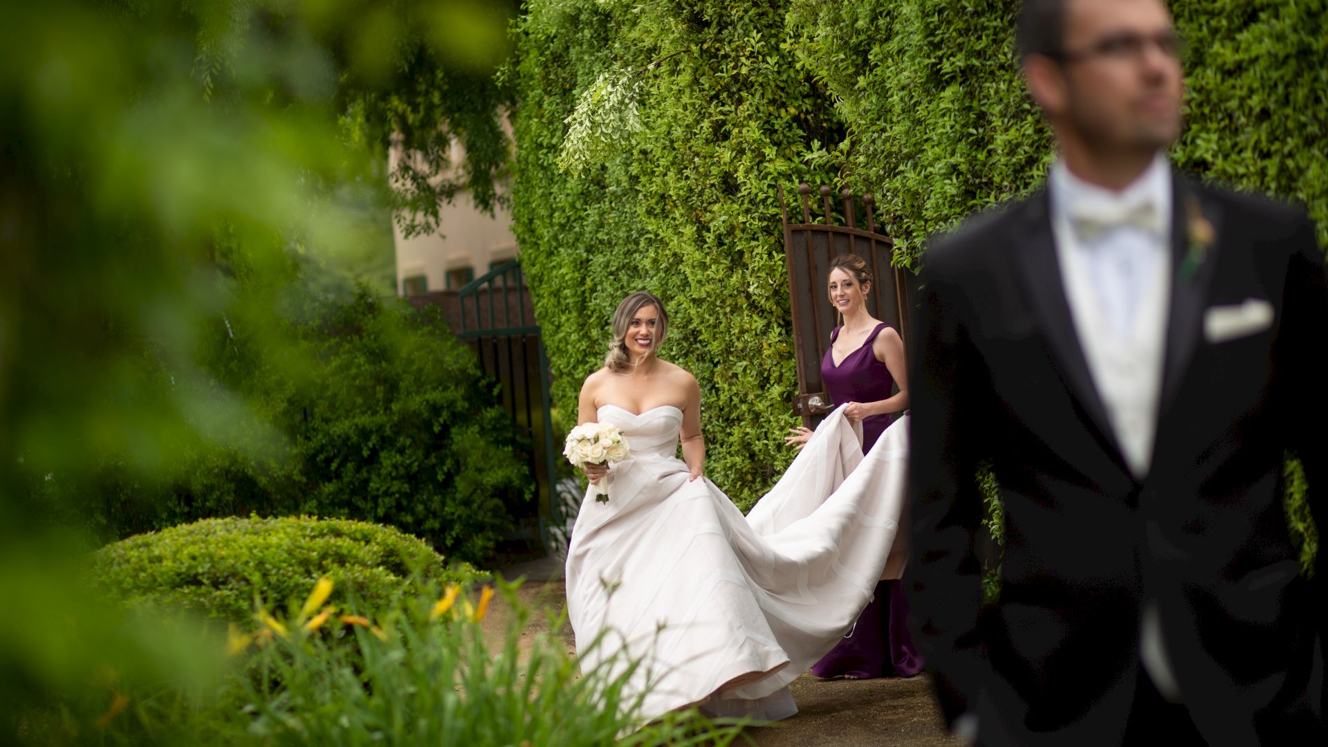 A bride holding a bouquet is walking with her bridesmaid in a garden, while a man in a suit stands in the foreground.