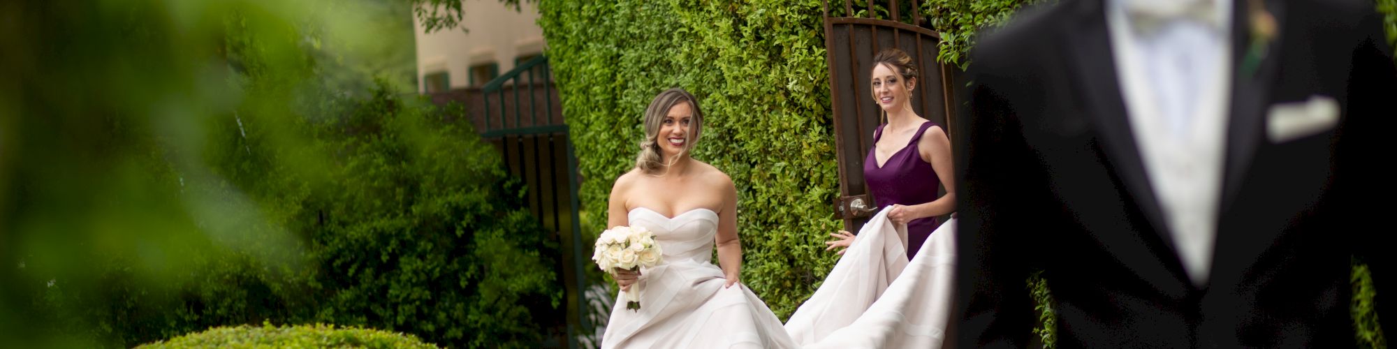 A bride holding a bouquet is walking with her bridesmaid in a garden, while a man in a suit stands in the foreground.