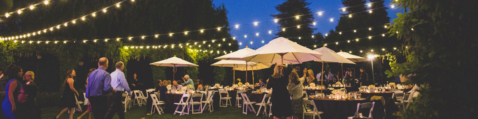 An outdoor evening event with people gathered under string lights and umbrellas, featuring tables set up on a lawn surrounded by trees and greenery.