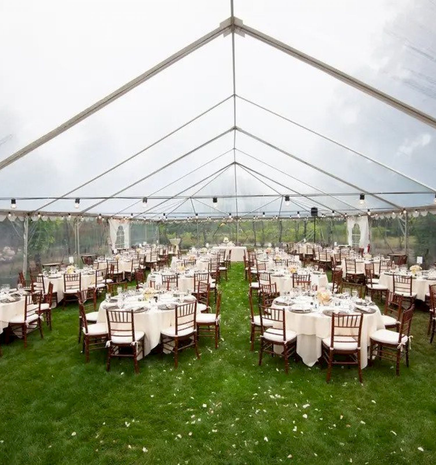 The image shows a large outdoor event setup under a transparent tent with multiple round tables and chairs arranged on a grassy area, prepared for guests.