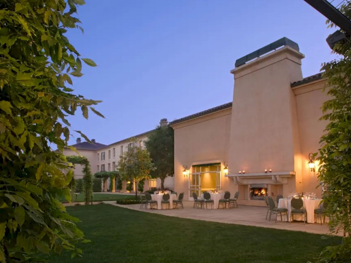 A courtyard featuring a well-lit building with an outdoor fireplace, surrounded by a neatly manicured lawn and various dining tables.