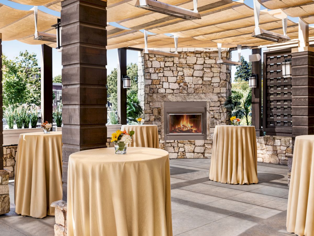 The image shows an outdoor setup with round, high tables covered in beige tablecloths, a stone fireplace, and overhead pergola.