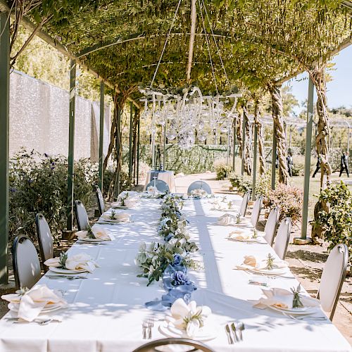 A long outdoor dining table set under a green canopy, decorated with white linens, greenery, and floral arrangements in a serene garden setting.