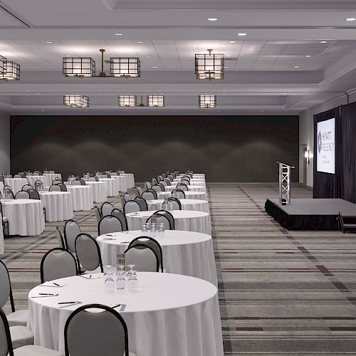 An empty conference room setup with round tables and chairs, a stage, and two screens displaying 