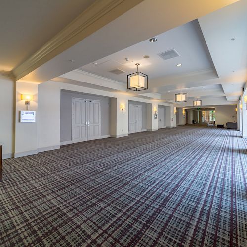 A spacious, well-lit hallway with patterned carpet, wooden chairs, white walls, and ceiling lights, leading to a glass door with a view outside.