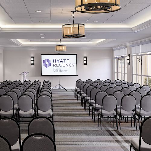 A conference room setup with rows of chairs facing a screen displaying the Hyatt Regency logo, with windows and ceiling lights.