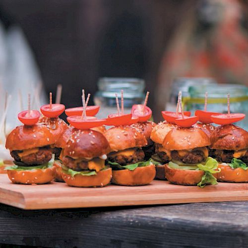 The image shows a serving tray with mini sliders each topped with a small tomato half, with additional appetizers in the background.