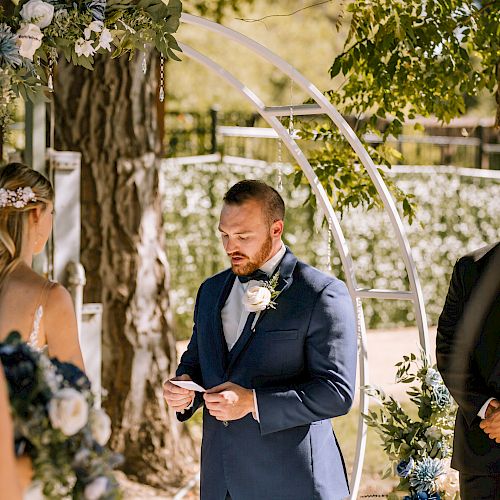 A wedding ceremony with a man in a blue suit reading from a paper, surrounded by three other people, and floral decorations in the background.