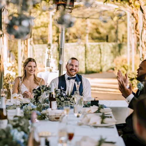 A couple sits at a decorated table, smiling and talking with guests in an outdoor setting with a warm, festive atmosphere, possibly at a wedding.