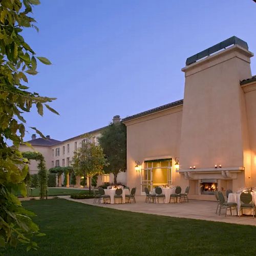 An elegant outdoor area featuring a large building with a patio, tables, chairs, ambient lighting, greenery, and a fireplace at dusk.