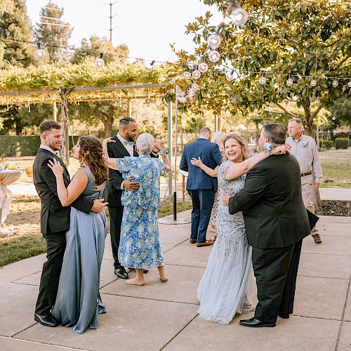 People in formal attire are dancing outdoors at a summer event, surrounded by greenery and string lights on a sunny day.