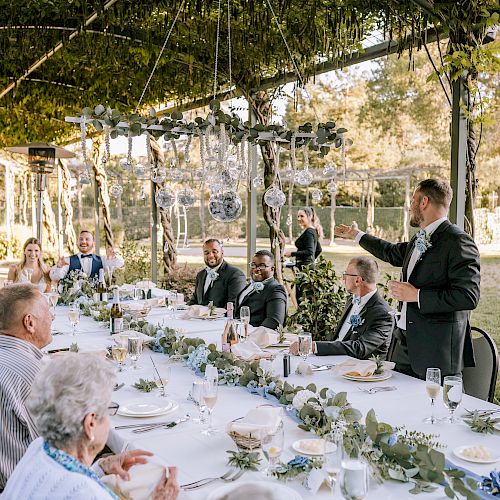 A gathering of people seated at a long, decorated table outdoors, listening to a man in a suit who is giving a speech, with nature in the background.