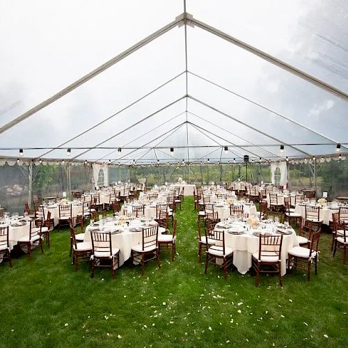 A large outdoor event tent with numerous round tables set with white tablecloths and chairs, arranged on a green lawn, prepared for a gathering.