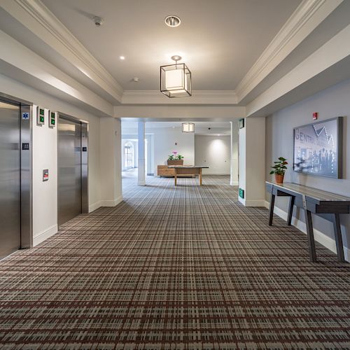 A hallway with plaid carpet, two elevators on the left, a table with plants on the right, and another table at the far end under a window.