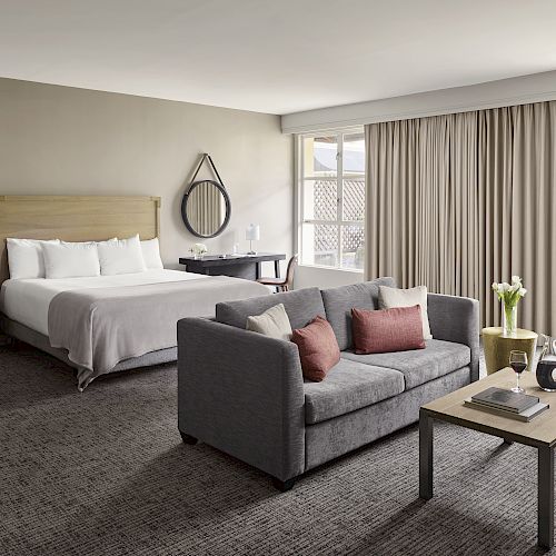 A spacious hotel room with a king-sized bed, a gray sofa, armchair, coffee table, and a work desk by the window. Neutral tones dominate the decor.