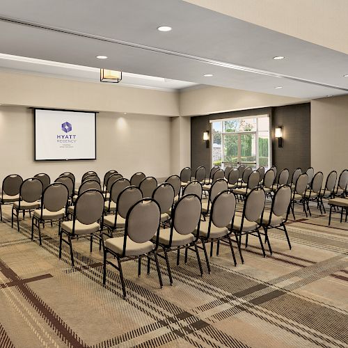 The image shows an empty conference room with rows of chairs facing a projector screen and a podium. The room has patterned carpeting.