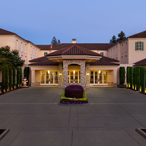 The image shows a grand building with a driveway leading to an entrance, flanked by tall trees, with a manicured garden in the center.